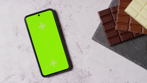 person picking up green screen mobile phone next to bars of milk plain and white chocolate