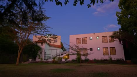 Volando-A-Través-De-Los-árboles-Sobre-El-Jardín-Verde-Hacia-La-Casa-Unifamiliar-Al-Atardecer