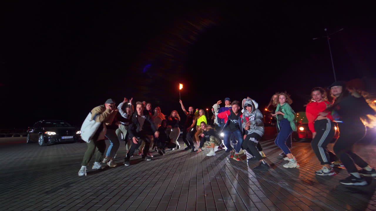 Hip hop dancers at the night square. Man holding a burning torch standing in the middle. Cars with headlights on at backdrop.