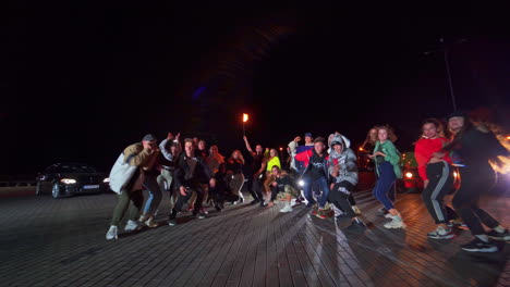 hip hop dancers at the night square. man holding a burning torch standing in the middle. cars with headlights on at backdrop.