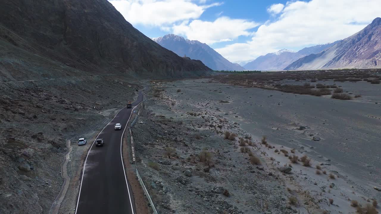 Aerial drone shot capturing a vehicle navigating through the diverse terrain of Nubra Valley, from desert plains to green patches.