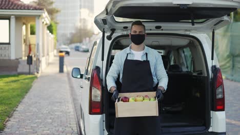 hombre amigable sonriente con máscara sosteniendo una caja de madera con manzanas en la calle al aire libre, retrato de hombre de entrega feliz en delantal y