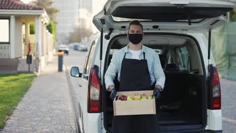 friendly smiling man in mask holding wooden box with apples at street outdoors, portrait of happy delivery man in apron and