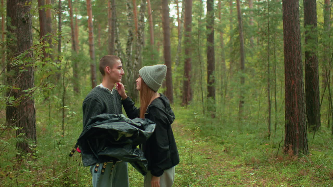 Lady gently removes dirt from cousin eye while standing in forest, playfully adjusting beanie he pulled over her face, both smiling in lighthearted moment surrounded by tall trees and camping gear