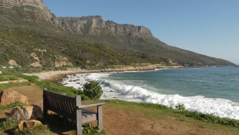 Un-Banco-Solitario-Domina-La-Playa-Rocosa-Y-Las-Montañas-En-Ciudad-Del-Cabo,-Rsa