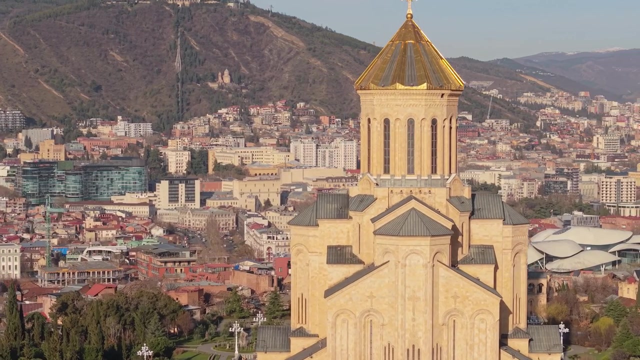Záběr dronem odhalující Katedrálu Nejsvětější Trojice v Tbilisi s její zlatou kupolí v teplém ranním světle a jemných atmosférických tónech.
