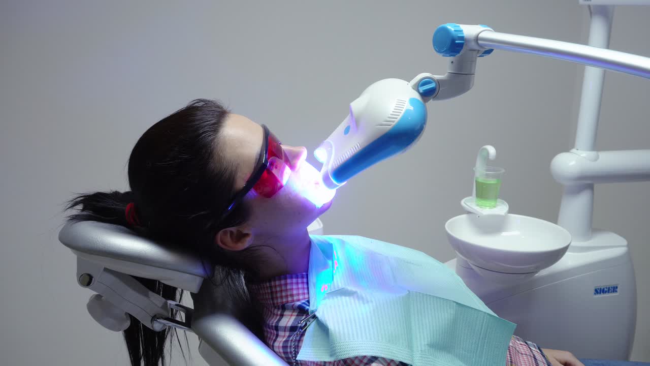 Young woman with an expander in mouth and red protective glasses getting UV whitening at the dentist's office. An ultra violet whitening machine in operation on a patient's teeth