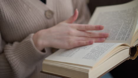 primer plano de una mano delicada con uñas bien cuidadas volviendo suavemente las páginas de un libro abierto, con ropa tejida, fondo ligeramente borroso