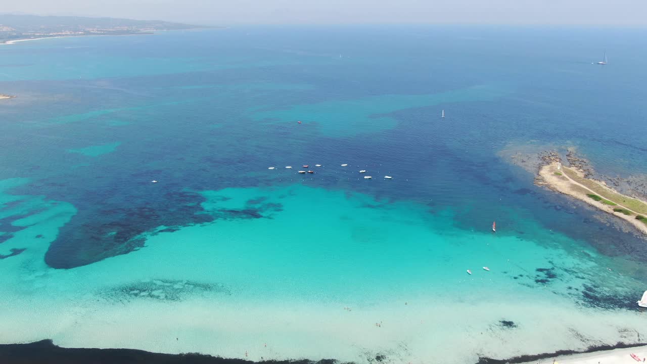 This video follows a forward and downward motion of white sandy shoreline and crystal clear water with boats and sails