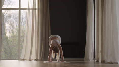 A-young-woman-in-white-sportswear-is-stretching-with-a-large-hall-with-large-windows-in-a-slow-motion-scheme-the-sun's-rays-shine-through-the-window.-Healthy-lifestyle-healthy-morning