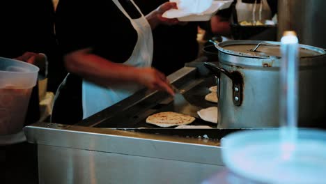 woman flipping several mexican tortilla on a grill. huge pot in the foreground with beans cooking inside. customers waiting for the food to be placed in a box.