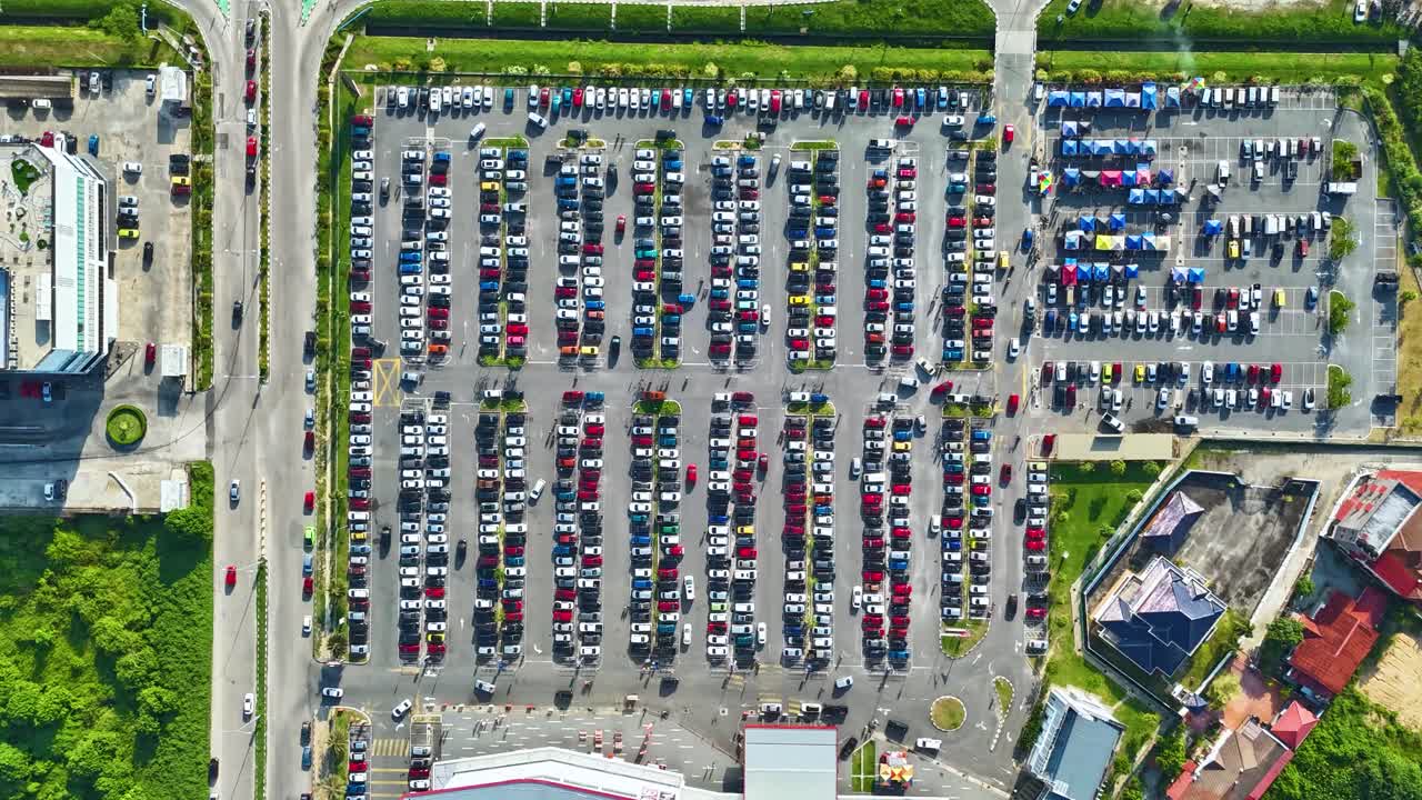 Top-down drone hyperlapse of a super busy car park in Kota Bharu, Malaysia on a weekend. Rows of colorful vehicles and shoppers’ cars fill the parking area near a shopping complex