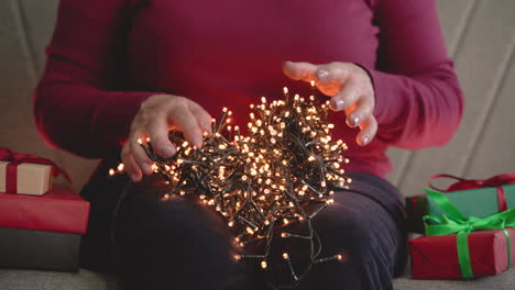 vista de cerca de las manos de una mujer sosteniendo luces de navidad sentadas en el sofá con regalos de navidad alrededor