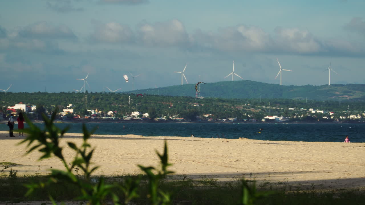 풍력 에너지 농장이 배경으로 운영되고 해변에서 카이트서핑을 즐기고 사진을 찍는 방문객들이 있는 개발된 교외의 해변 전망