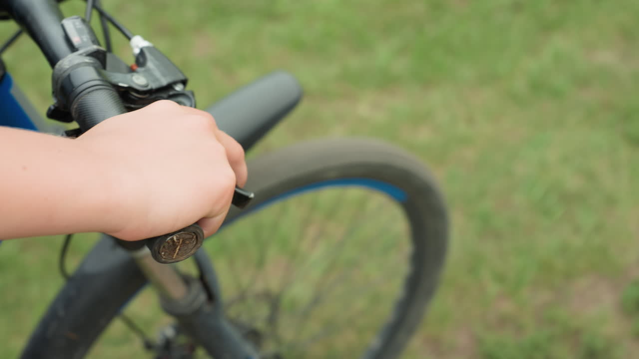 Primer plano del puño del manillar y la rueda delantera, primer plano de una mano accionando la maneta de freno de una bicicleta de montaña azul, parque cubierto de hierba al fondo, movimiento sutil que indica el inicio del paseo, enfoque en la textura del puño