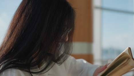 side view of cheerful lady in glasses smiling and reacting to novel she is reading, dressed in white shirt, standing near wooden rail in bright interior with glass windows and warm natural light