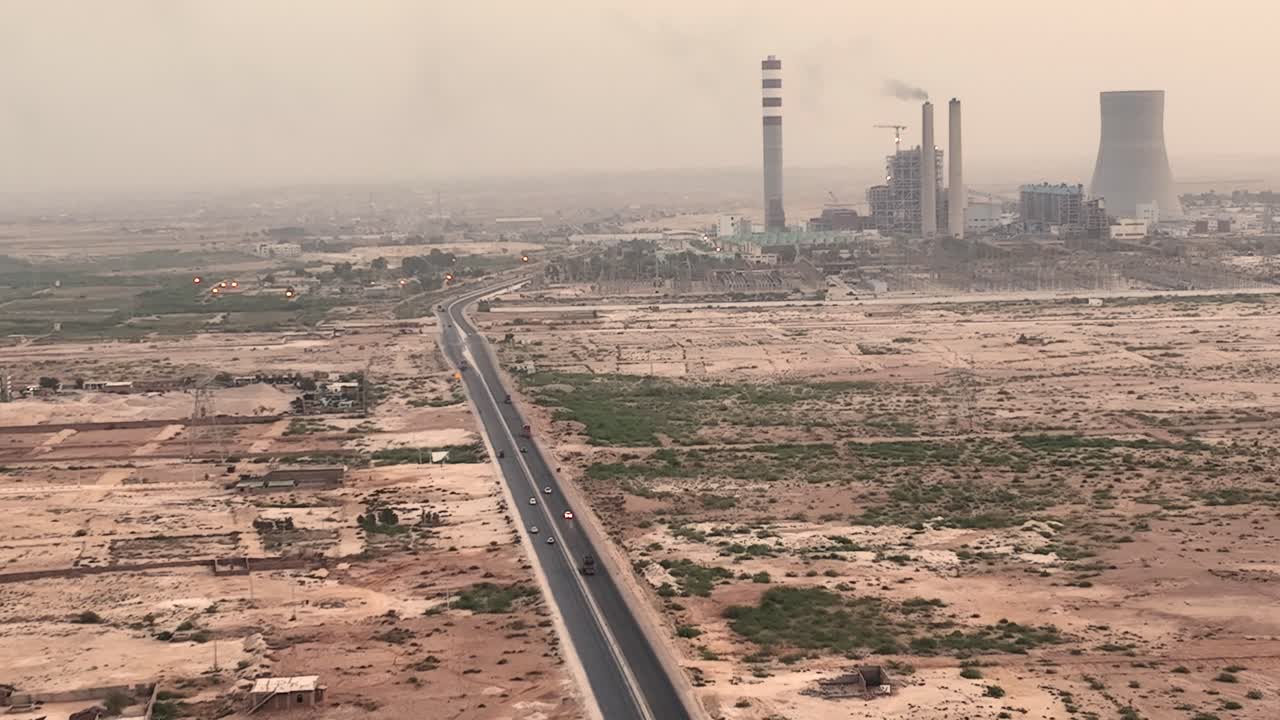toma aérea cinematográfica graduada en color de un coche plateado conduciendo por una carretera rural en el desierto y grandes chimeneas en un fondo lejano