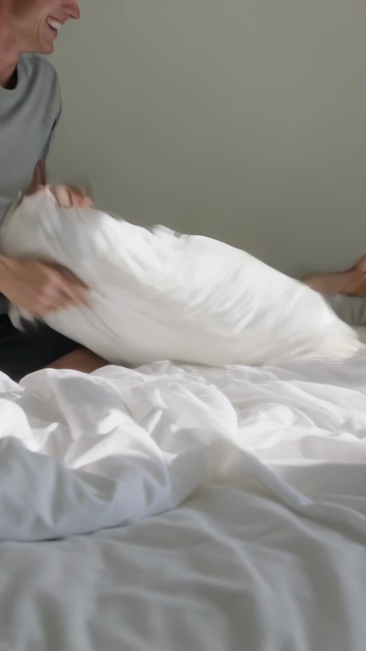 Video vertikal: Pria berbaju abu-abu dan celana pendek hitam meraih, mengayunkan bantal melintasi tempat tidur, untuk bermain