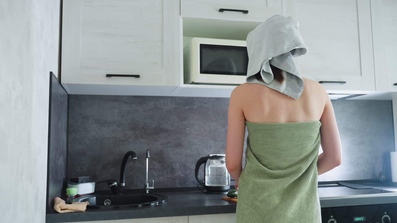 Back view of woman wrapped in green towel standing in well-furnished kitchen, she is enjoying a moment of relaxation in a modern kitchen setting, surrounded by contemporary appliances