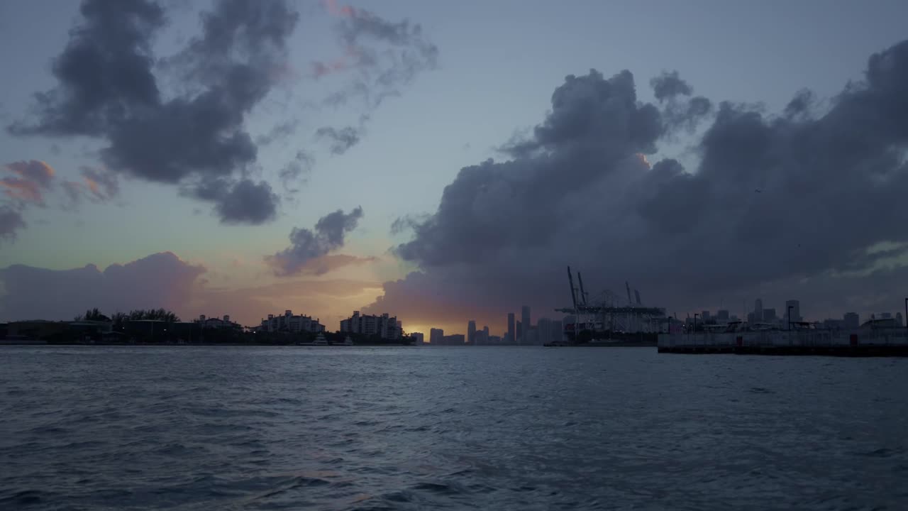 Twilight descends over downtown Miami as cargo cranes and towers silhouette against a fiery ocean sunset.