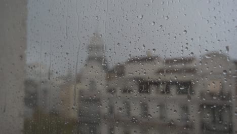 a slow-motion close-up view of heavy raindrops seen through a window, with an urban city landscape in the background