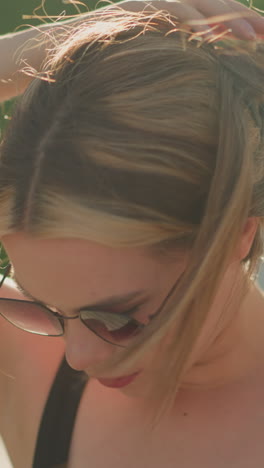 dama ajusta su cabello como el viento sopla a través de él mientras usa gafas de sol al aire libre, el sol irradia en ella, y un fondo de vegetación borrosa
