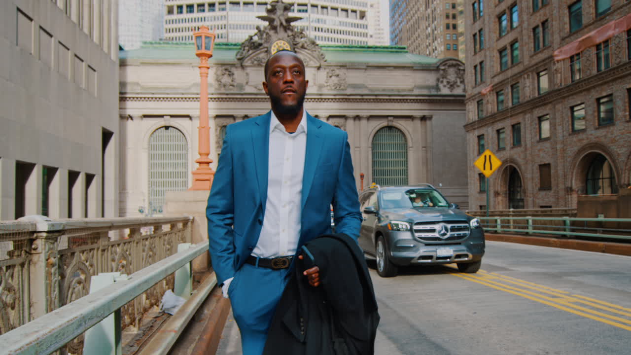 New York, USA, 1 aprile 2026: Uomo d'affari che cammina vicino al Grand Central Terminal a Manhattan. Uomo d'affari di colore in abito blu che cammina per strada vicino al Grand Central Terminal a New York, tenendo un cappotto.