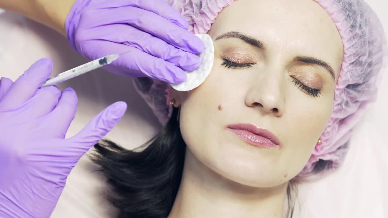 Patient in medical cap receives facial injection by the specialist in beauty salon. Procedure of face aging injection