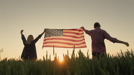 una pareja feliz levanta la bandera de los estados unidos, de pie contra el telón de fondo de un campo de trigo al atardecer.