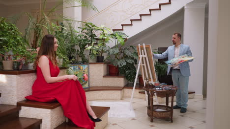 artist creates painting of pretty woman in residence. beautiful mansion owner in red dress with rose performs posture for art master in luxury house