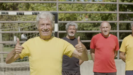 vorderansicht eines glücklichen senior-fußballspielers, der in die kamera blickt und auf dem fußballplatz eine geste mit dem daumen nach oben zeigt