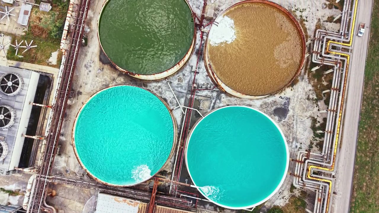 Large round tanks filled with colored water are seen from above in an industrial area during the day