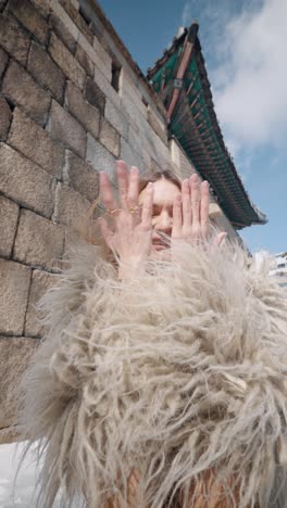 happy smiling shy blond woman model cover her face with hands wearing winter fur jacket looking directly at camera with gwanghuimun gate historical landmark of seoul city in background, south korea
