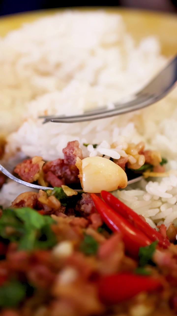 Secuencia en primer plano de una cuchara y un tenedor recogiendo arroz, huevo cocido y salchicha de cerdo fermentada de un colorido plato del norte de Tailandia bajo una cálida iluminación interior