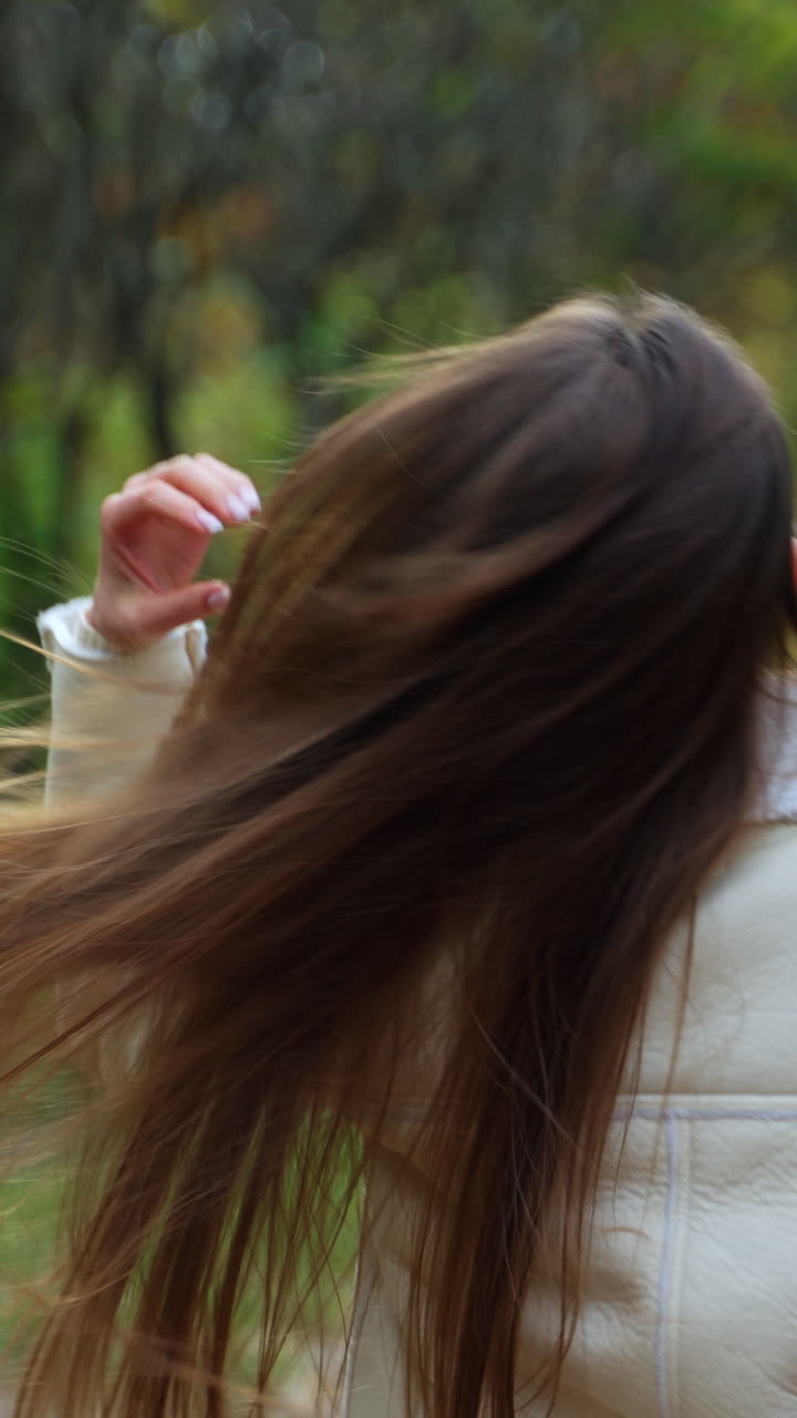 Rear view of a brunette girl walking by the park in autumn. Lady turns around for a moment showing a cup in her hand. Blurred backdrop. Vertical video