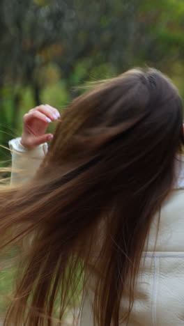 rear view of a brunette girl walking by the park in autumn. lady turns around for a moment showing a cup in her hand. blurred backdrop. vertical video