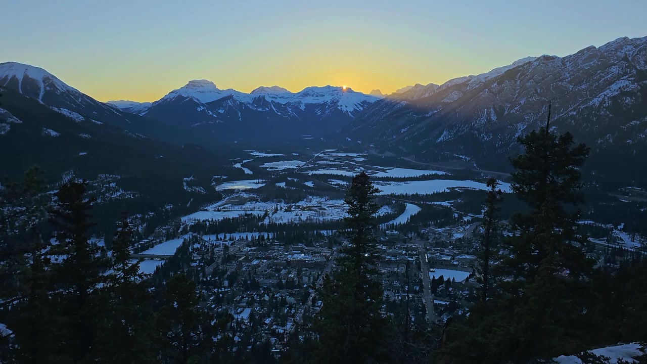 Mặt trời lặn trên Banff, Alberta, với Thung lũng Bow phủ tuyết, thị trấn miền núi bên dưới, những bóng cây thường xanh sẫm màu, đường chân trời rực rỡ, những khúc sông đóng băng uốn lượn và khung cảnh mùa đông hùng vĩ của dãy Rockies