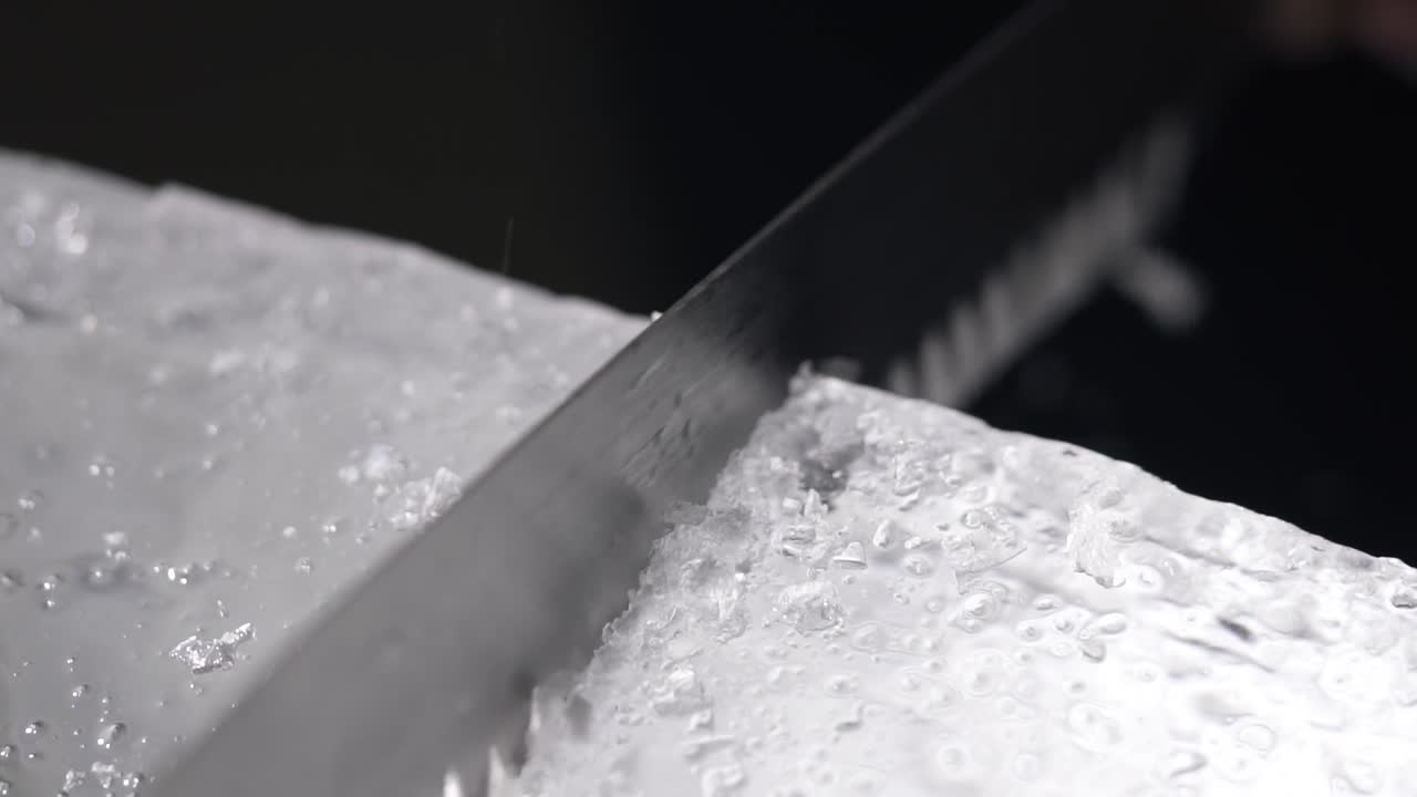 A bartender is cutting the ice block to smaller pieces for cocktail service by using a long toothed knife.