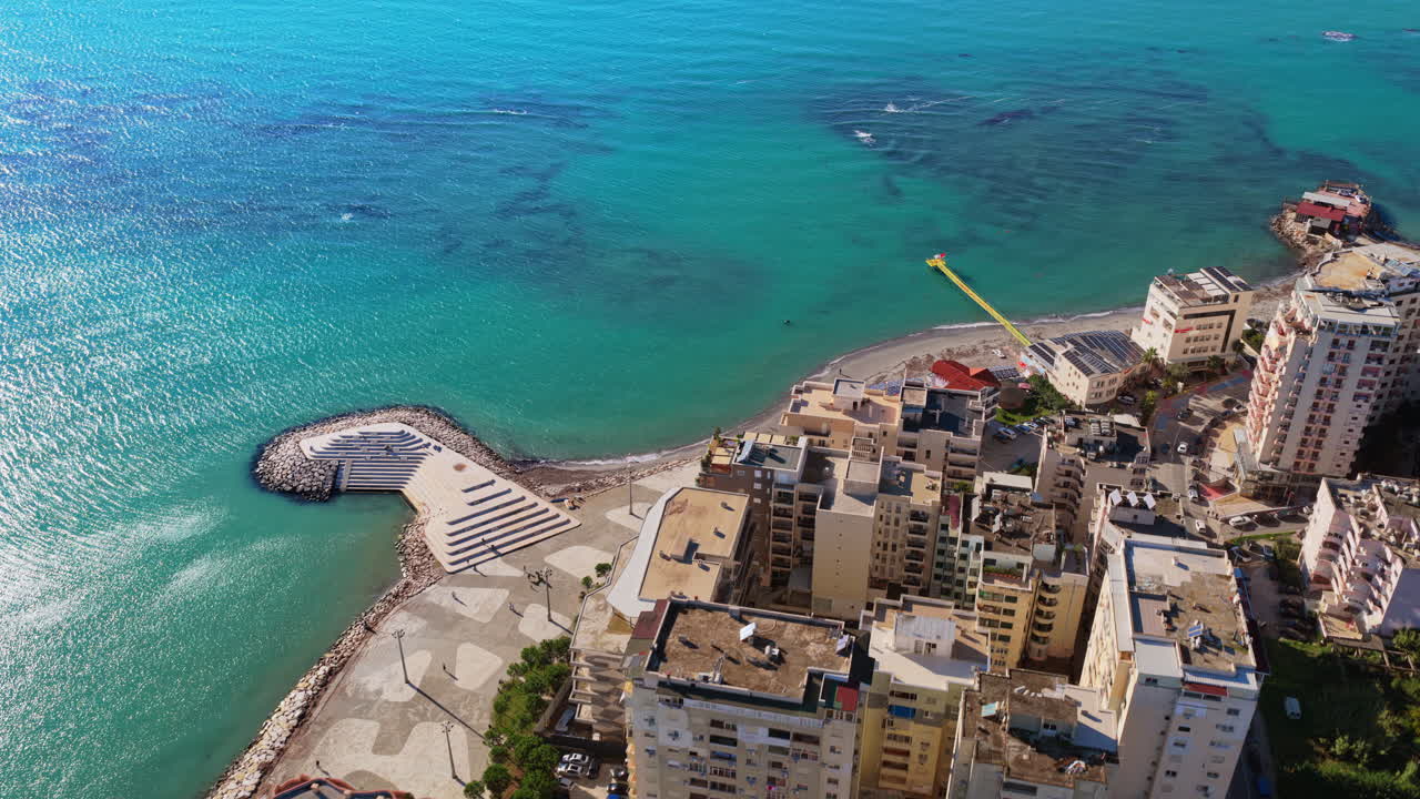Vista aerea da drone della costa di Durazzo che mostra alti palazzi, mare turchese e la piattaforma sul lungomare dalla forma unica che si estende nell'acqua