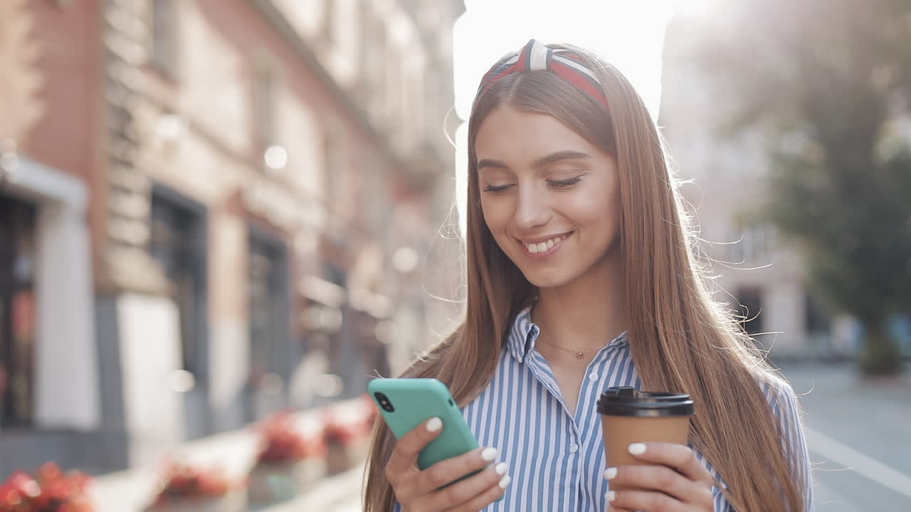 Giovane donna che sorride mentre usa lo smartphone e tiene un caffè in una via cittadina.