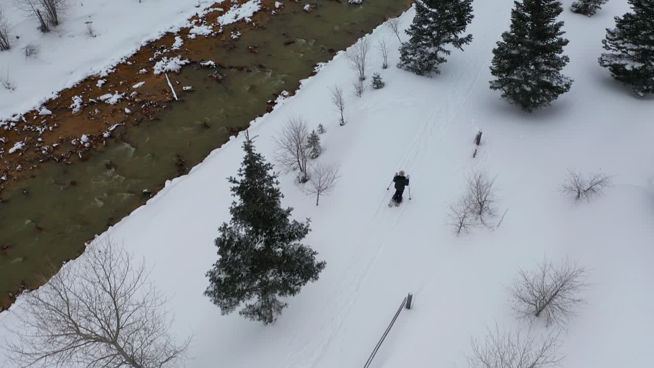 공중 전망, 겨울 아침 풍경에서 강가에서 눈 위를 걷고 있는 스노슈를 입은 여성, 드론 샷을 기울입니다.