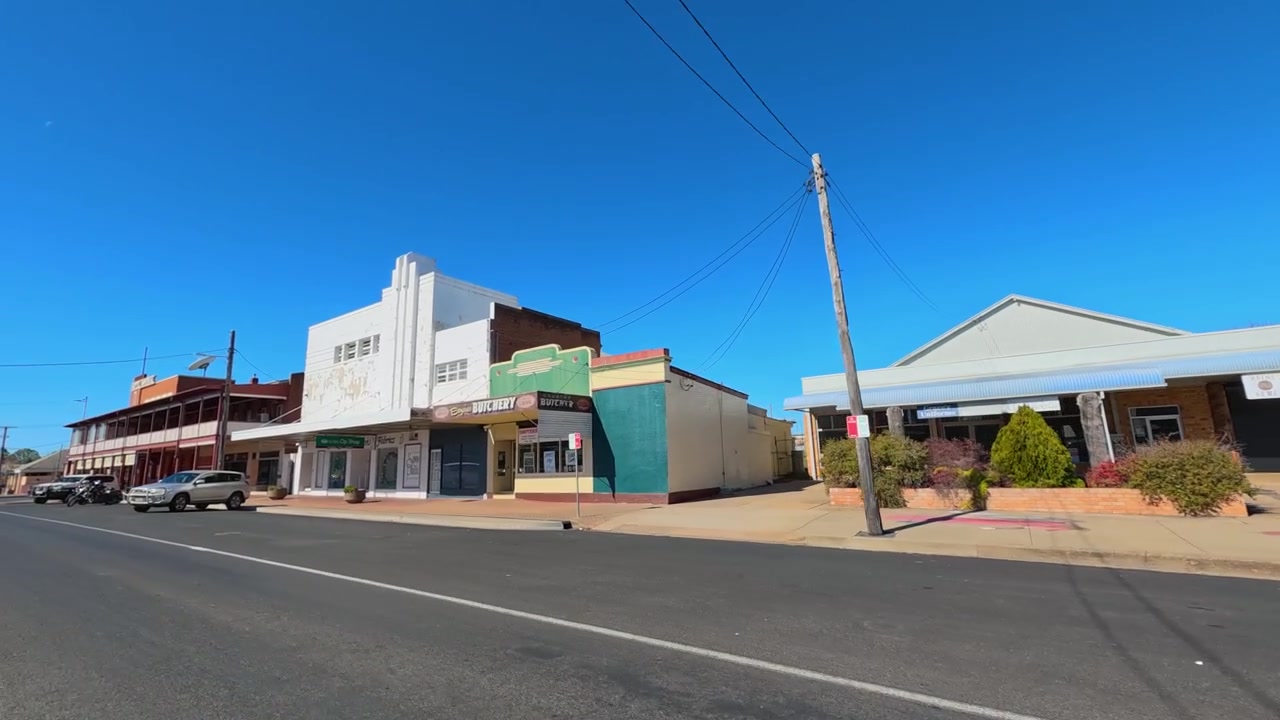Vehículo circulando pasando edificios rurales históricos bajo un cielo azul claro en Coonabarabran