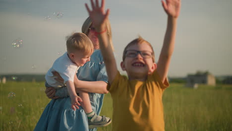 una mujer alegre en un vestido azul sostiene a un niño pequeño en una camisa blanca mientras un niño joven de amarillo se extiende emocionado a su lado. todos se deleitan tocando burbujas coloridas flotando alrededor