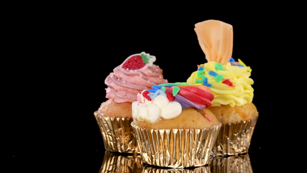 Three colorful gourmet cupcakes rotate on black background under bright studio lighting, close-up view