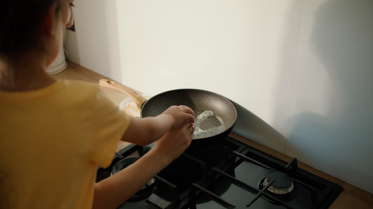 그녀의 어머니와 함께 노란색 드레스를 입은 작은 갈색 소녀의 뒷면은 고기 달을 프라이팬으로 부러뜨리고 현대 부에서 함께 긴 달을 준비합니다.