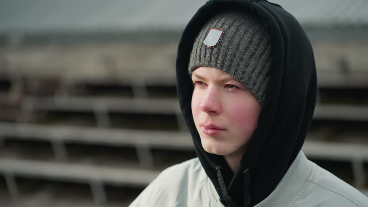 Close-up of a young boy wearing a gray sweater and black beanie, looking focused with a serious expression, the background is blurred, showing a staircase