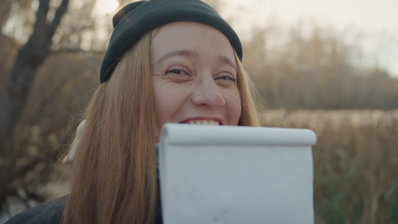 Donna bianca all'aperto con un blocco note, si nasconde dietro un album da disegno e sbircia con un sorriso timido, indossa un berretto e una sciarpa, ritratto spontaneo ravvicinato in una palude autunnale, luce dorata soffusa, canne e acqua.