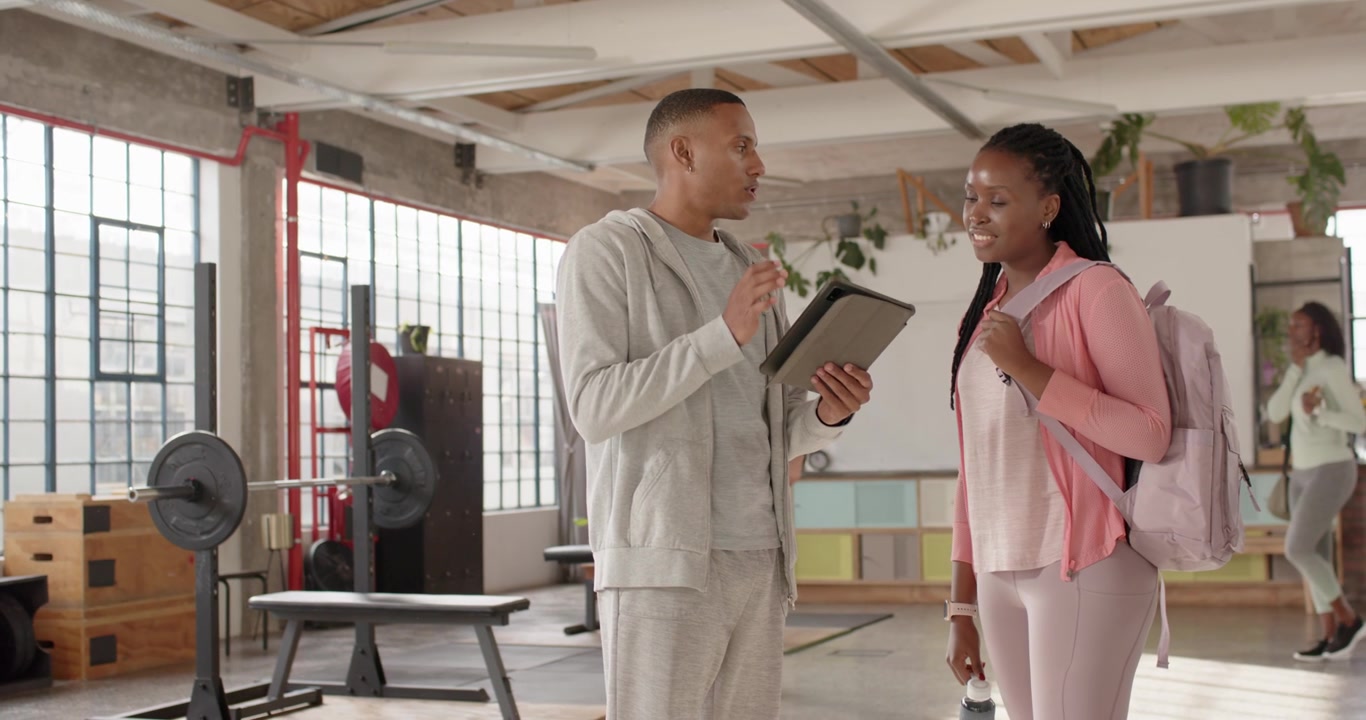 Male coach scrolling tablet explaining plan while female client approaching with pink pack at gym