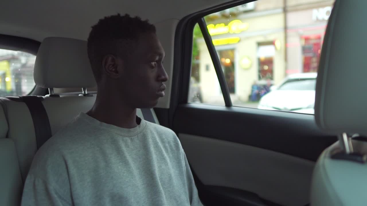 Handsome young black man rides on back seat of a car, looks around in wonder out of the window. Big city view reflected in window. Wearing white casual shirt. Slow motion