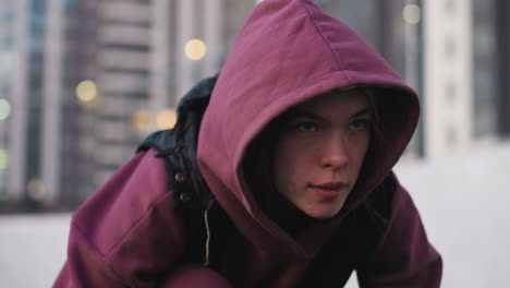 close up of female exercise lover kneeling with one leg and two hands down on black asphalt sports court near white barrier topped with chain link fence wearing maroon hoodie and white sneakers